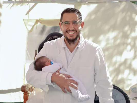 המוהל איתמר חמו בחלוק לבן מחזיק תינוק ומחייך