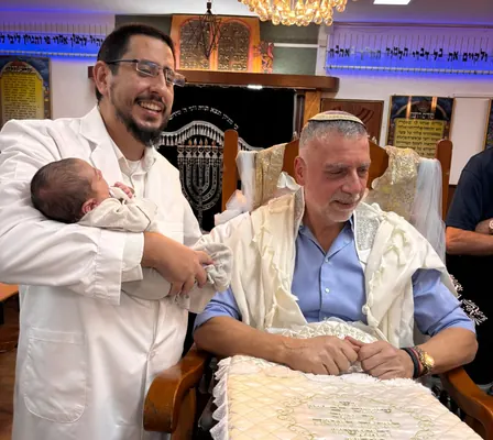 איתמר חמו מחזיק תינוק ליד הסנדק המחייך בבית הכנסת